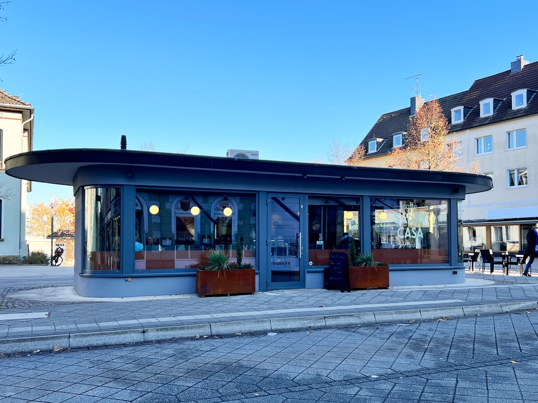 Außenansicht, "Caffé Santa Casa" in Heiligenhaus Ein modernes, rundes Café mit großen Fenstern und hölzernen Pflanzkästen vor der Fassade.