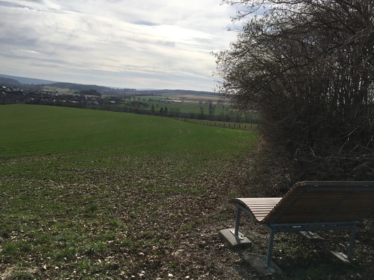 Aussicht auf Drenke Holzbank am Rand eines grünen Feldes mit weitem Blick auf die sanft geschwungene Landschaft.