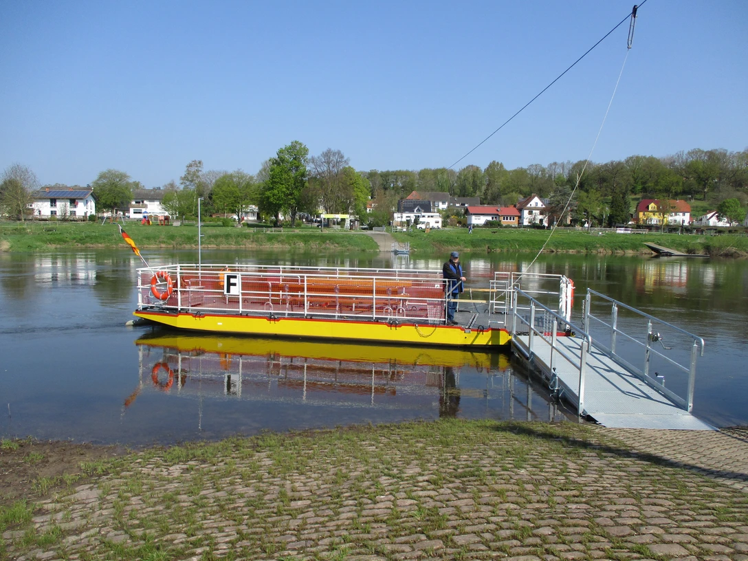 Eine gelbe Seilfähre überquert die Weser, im Hintergrund eine Kulisse aus Bäumen und Häusern.