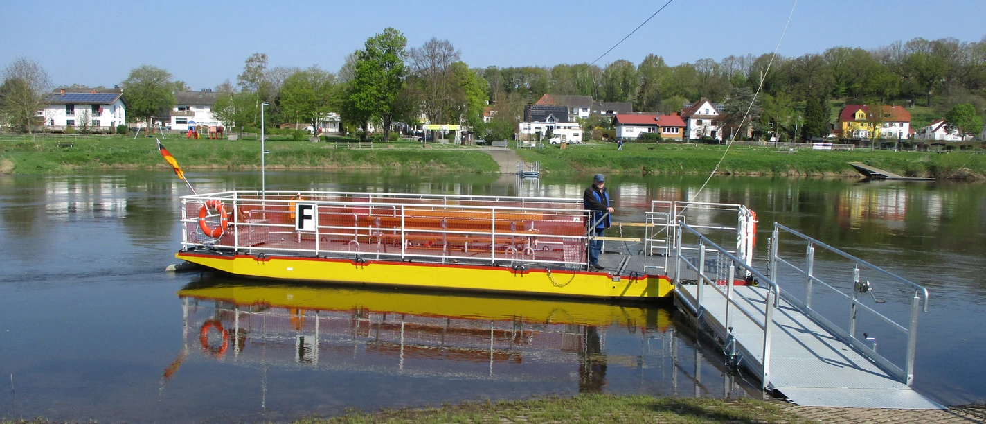Dreiländereckfähre Herstelle/Würgassen Eine gelbe Seilfähre überquert die Weser, im Hintergrund eine Kulisse aus Bäumen und Häusern.