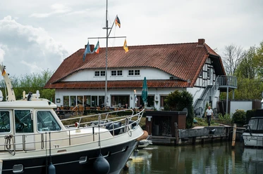Yachthafen Lübbecke Weiße Motorjacht im Hafen vor einem Gebäude mit rotem Ziegeldach und Fähnchen, bewölkter Himmel.