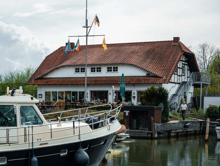 Yachthafen Lübbecke Weiße Motorjacht im Hafen vor einem Gebäude mit rotem Ziegeldach und Fähnchen, bewölkter Himmel.