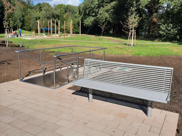 Kurpark Levern Silberne Sitzbank mit barrierefreiem Zugang auf gepflasterter Fläche, umgeben von grünem Parkgelände.