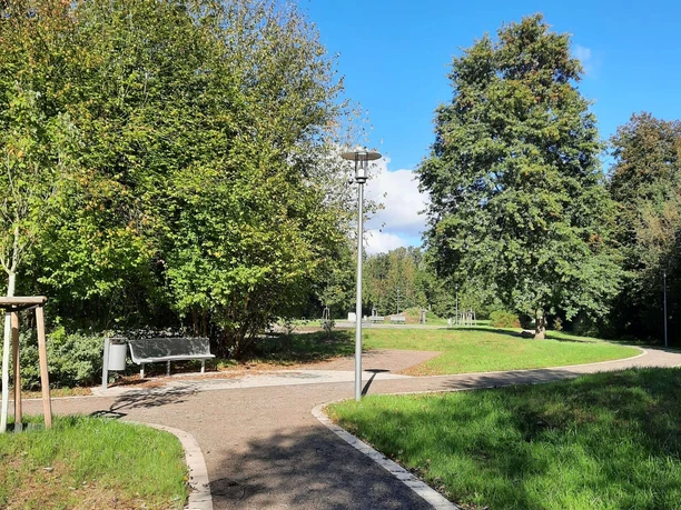Parkweg mit Bäumen und Bänken unter blauem Himmel, von Laternen entlang des Weges gesäumt.