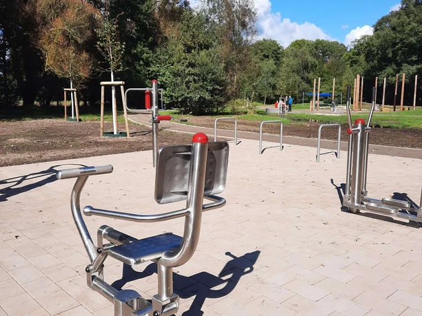 Silberne Outdoor-Fitnessgeräte auf gepflastertem Areal in einem grünen Park bei Sonnenwetter.