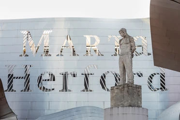 Ein modernes Gebäude mit einer markanten Metallfassade, davor eine Skulptur eines stehenden Mannes.