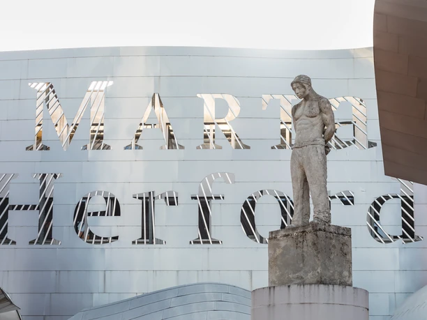Ein modernes Gebäude mit einer markanten Metallfassade, davor eine Skulptur eines stehenden Mannes.