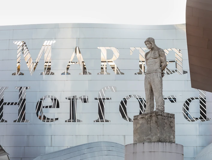 Ein modernes Gebäude mit einer markanten Metallfassade, davor eine Skulptur eines stehenden Mannes.