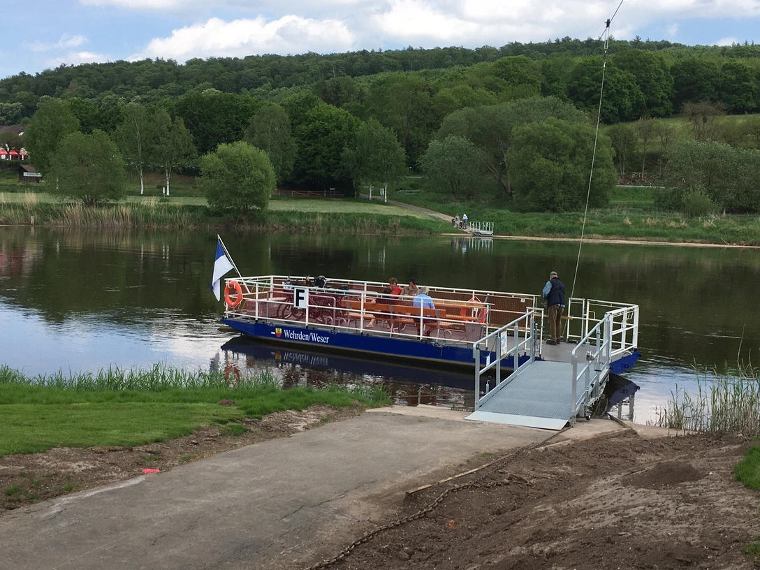 Personenfähre "Weser-Waler" dockt auf ruhigem Fluss an, umgeben von üppigem Grün und Wanderwegen.