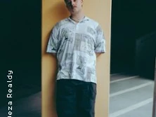 Ein Mensch in lässiger Kleidung lehnt an einer orangefarbenen Wand, umgeben von dunkler Umgebung.