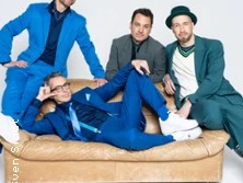 Vier Männer in moderner, bunter Kleidung posieren stilvoll auf einem braunen Sofa vor einem hellen Hintergrund.