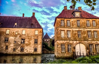schloss_tatenhausen_torgebaeude.jpg Historisches Wasserschloss mit zwei turmähnlichen Gebäuden, umgeben von einem Wassergraben im Abendlicht.