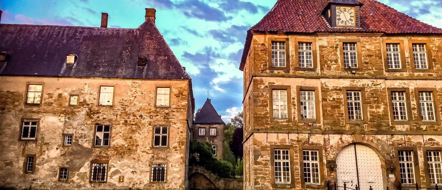 schloss_tatenhausen_torgebaeude.jpg Historisches Wasserschloss mit zwei turmähnlichen Gebäuden, umgeben von einem Wassergraben im Abendlicht.
