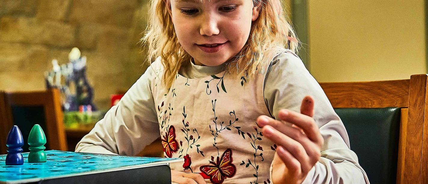 Spielewochen in der Sächsischen Schweiz Ein blondes Mädchen wirft lächelnd einen Würfel, während es an einem Tisch in einem Spiel vertieft ist.