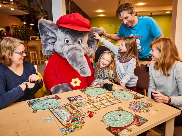 Gezinnen spelen bordspelletjes met Benjamin de olifant in het pretpark; lachende gezichten.