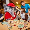 Benjamin Blümchen Spieletag Familien spielen begeistert Brettspiele mit Benjamin Blümchen im Freizeitpark; lächelnde Gesichter.Families play board games with Benjamin the Elephant in the amusement park; smiling faces.Rodiny hrají v zábavním parku stolní hry se slonem Benjaminem; usměvavé tváře.Rodziny grają w gry planszowe ze słoniem Benjaminem w parku rozrywki; uśmiechnięte twarze.Gezinnen spelen bordspelletjes met Benjamin de olifant in het pretpark; lachende gezichten.Famiglie che giocano a giochi da tavolo con Benjamin l'elefante nel parco divertimenti; volti sorridenti.