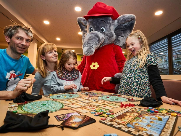 Gezin speelt bordspel met lachende Benjamin de olifant in pretpark, vrolijke sfeer.