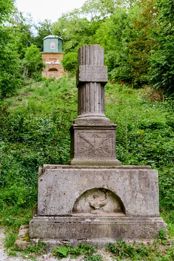 Kaffeemühle Eine antike Säule steht in einer grünen Parklandschaft, umgeben von Bäumen und einem steinernen Weg.