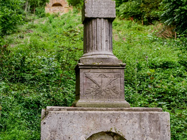 Kaffeemühle Eine antike Säule steht in einer grünen Parklandschaft, umgeben von Bäumen und einem steinernen Weg.