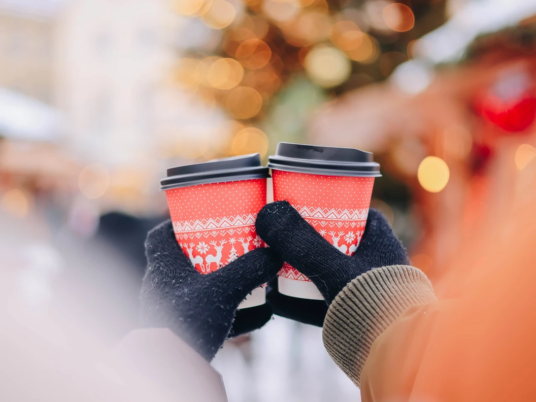 Wintermarkt Greetsiel Zu sehen sind zwei Hände mit Handschuhen an, die zwei Glühweinbecher in der Hand halten. Im Hintergrund erkennt man verschwommen die Umrisse eines geschmückten Tannenbaums.