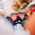 Wintermarkt Greetsiel Zu sehen sind zwei Hände mit Handschuhen an, die zwei Glühweinbecher in der Hand halten. Im Hintergrund erkennt man verschwommen die Umrisse eines geschmückten Tannenbaums.