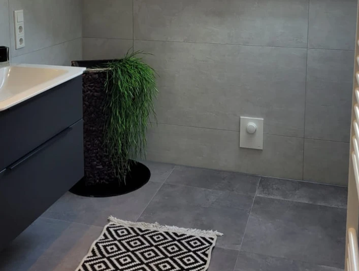 Modernes Badezimmer mit grauen Fliesen, schwarzem Waschbeckenunterschrank und dekorativem Teppich.