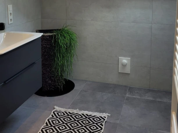 FeWo_Brinkmann Stukenbrock Senne © L_Brinkmann (2).jpg Modernes Badezimmer mit grauen Fliesen, schwarzem Waschbeckenunterschrank und dekorativem Teppich.