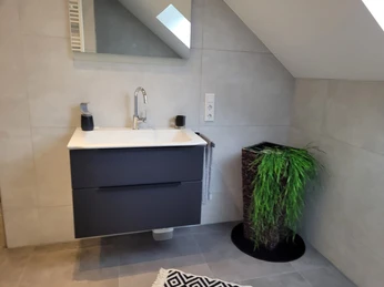 Badezimmer mit modernem Waschbecken, großen grauen Fliesen und einer Pflanze in einem geflochtenen Korb.