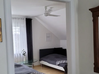 Ein helles Zimmer mit zwei Einzelbetten, Vorhängen, einem Glastisch und einem Deckenventilator.