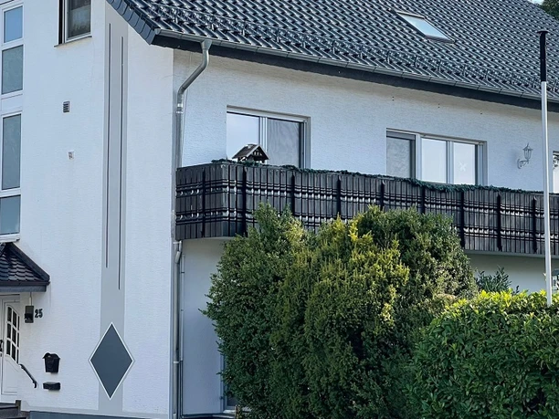 Ein weißes, zweistöckiges Gebäude mit schwarzen Balkonen und Dach, umgeben von grünen Büschen.