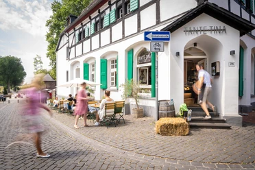 Außenbereich Tante Tina Das Bild zeigt den Außenbereich von Tante Tina mit Gästen, die auf einer Kopfsteinpflasterstraße sitzen.