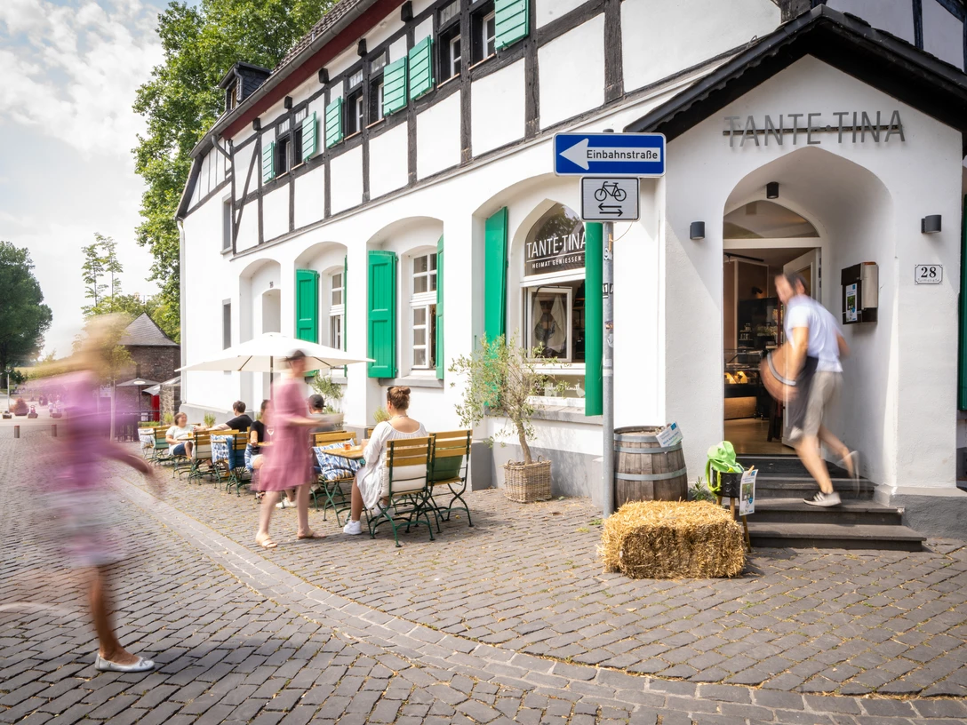 Außenbereich Tante Tina Das Bild zeigt den Außenbereich von Tante Tina mit Gästen, die auf einer Kopfsteinpflasterstraße sitzen.