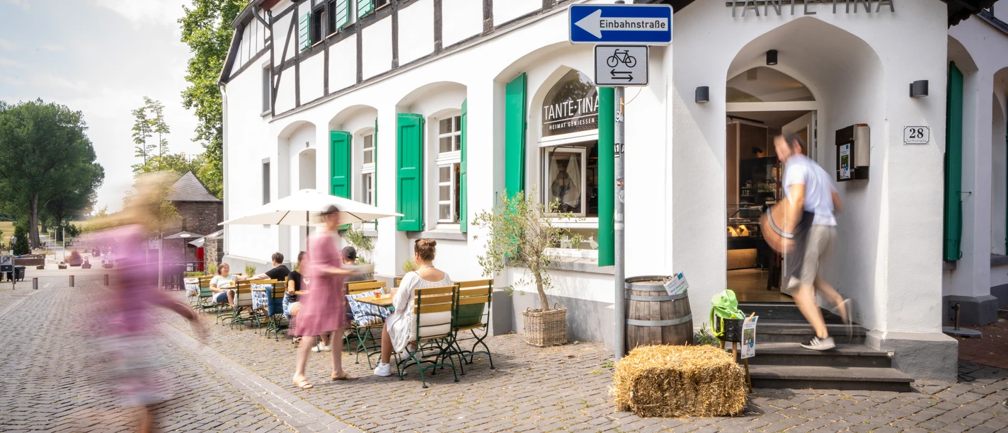 Außenbereich Tante Tina Das Bild zeigt den Außenbereich von Tante Tina mit Gästen, die auf einer Kopfsteinpflasterstraße sitzen.