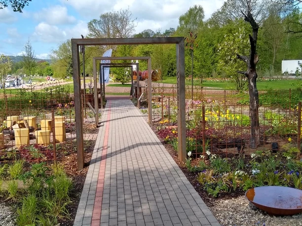 schoepfungsgarten_c_team_schoepfungsgarten (1).jpeg Gartenweg mit Blumenbeeten und Skulpturen, im Hintergrund Teich und Bäume an einem sonnigen Tag.