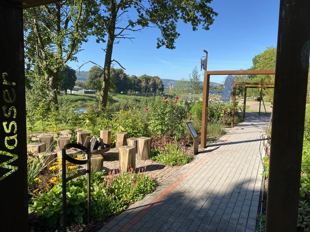 Grünanlage mit gepflastertem Weg, Pergola und Feuerschale mit Holzstamm Sitzhockern