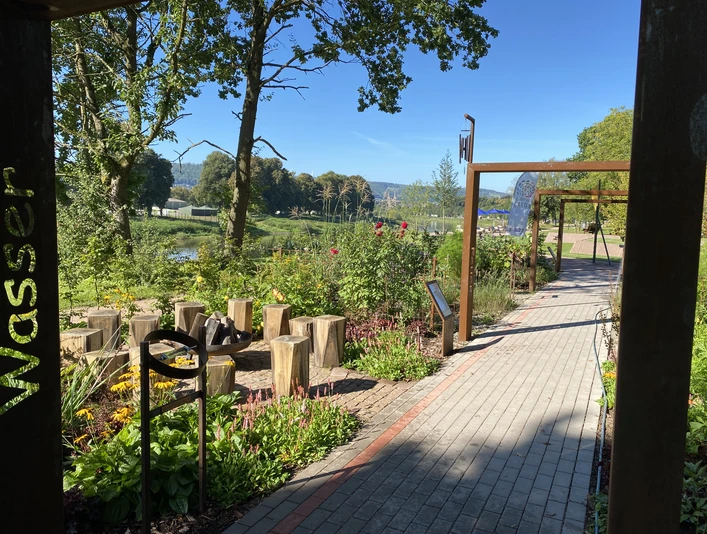 schoepfungsgarten_c_team_schoepfungsgarten (3).jpeg Grünanlage mit gepflastertem Weg, Pergola und Feuerschale mit Holzstamm Sitzhockern