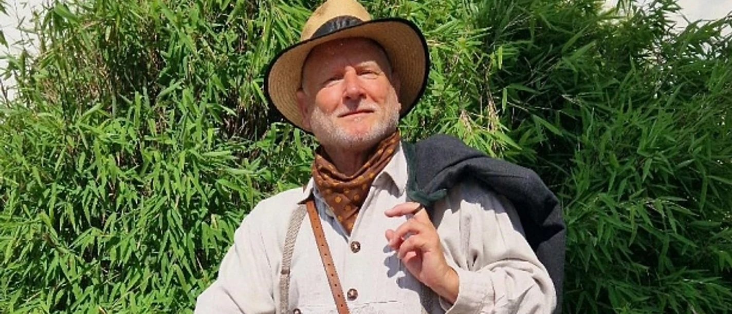 stadtfuehrer-900010141-22700-12.webp Älterer Mann in traditioneller Wanderkleidung mit Hut und Stock, umgeben von grüner Vegetation.