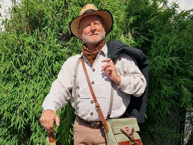 stadtfuehrer-900010141-22700-12.webp Älterer Mann in traditioneller Wanderkleidung mit Hut und Stock, umgeben von grüner Vegetation.