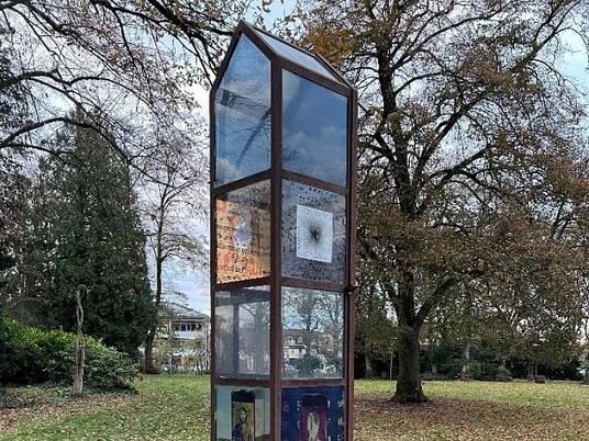 skulptur-900010145-22700-22.webp Metallische Skulptur im Park mit verzierten Glaspaneelen, umgeben von herbstlichen Bäumen und Laub.