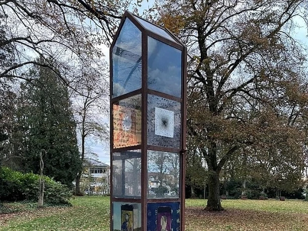 skulptur-900010145-22700-22.webp Metallische Skulptur im Park mit verzierten Glaspaneelen, umgeben von herbstlichen Bäumen und Laub.