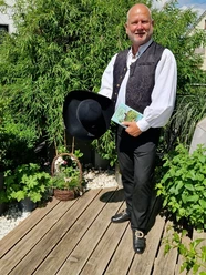 stadtfuehrer-900010142-22700-12.webp Ein lächelnder Mann in traditioneller Tracht steht auf einer Holzterrasse, umgeben von üppigem Grün.