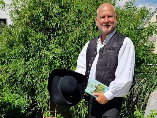 stadtfuehrer-900010142-22700-12.webp Ein lächelnder Mann in traditioneller Tracht steht auf einer Holzterrasse, umgeben von üppigem Grün.