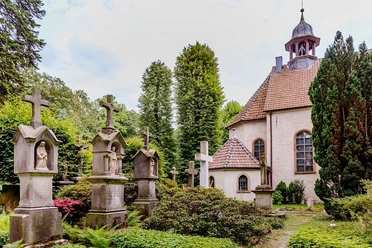 pfarrkirche_stockkaempen-900010262-22700-13.webp Historische Kapelle mit gepflegtem Friedhof, gesäumt von alten Grabsteinen und grüner Vegetation.
