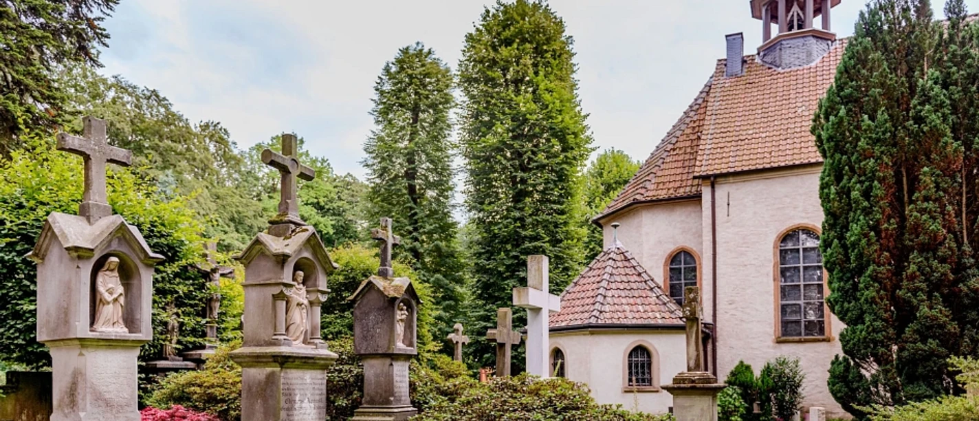 pfarrkirche_stockkaempen-900010262-22700-13.webp Historische Kapelle mit gepflegtem Friedhof, gesäumt von alten Grabsteinen und grüner Vegetation.