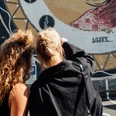 Mindwalk in Köln Zwei Frauen stehen im Sonnenlicht, schauen sich ein Kunstwerk auf einer Wand an. Die Parkplätze sind in der Nähe sichtbar, während die farbenfrohe kunstvolle Wand ein interessantes visuelles Motiv bietet.Two women stand in the sunlight, looking at a work of art on a wall. The parking spaces are visible nearby, while the colorful artistic wall provides an interesting visual motif.