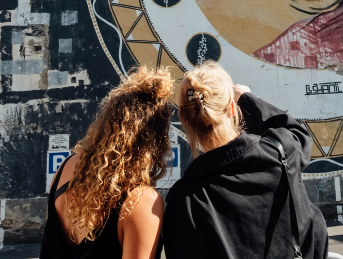 Mindwalk in Köln Zwei Frauen stehen im Sonnenlicht, schauen sich ein Kunstwerk auf einer Wand an. Die Parkplätze sind in der Nähe sichtbar, während die farbenfrohe kunstvolle Wand ein interessantes visuelles Motiv bietet.Two women stand in the sunlight, looking at a work of art on a wall. The parking spaces are visible nearby, while the colorful artistic wall provides an interesting visual motif.