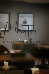 Blick in ein elegantes Restaurant mit dem Schriftzug "Matteo Pizza Pasta Vino" auf Glas.
