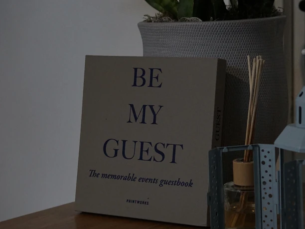 Tischdekor mit Gästebuch "Be My Guest", Pflanze in weißem Topf und Duftstäbchen auf Holzoberfläche.