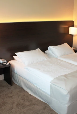 Superior twin room im relexa hotel Airport Düsseldorf-Ratingen.JPG Ein helles Hotelzimmer mit zwei Einzelbetten, zwei Nachttischen, Wandlampen und einem Sessel.