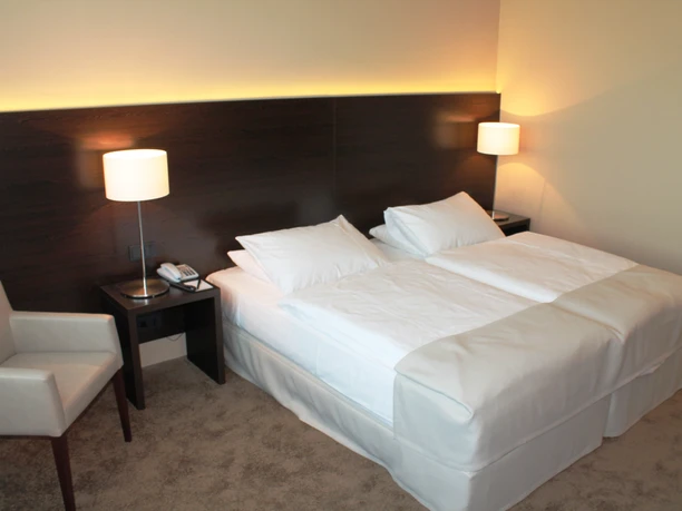 Superior twin room im relexa hotel Airport Düsseldorf-Ratingen.JPG Ein helles Hotelzimmer mit zwei Einzelbetten, zwei Nachttischen, Wandlampen und einem Sessel.